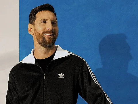 A smiling man in a black track jacket with white stripes, in front of a blue wall
